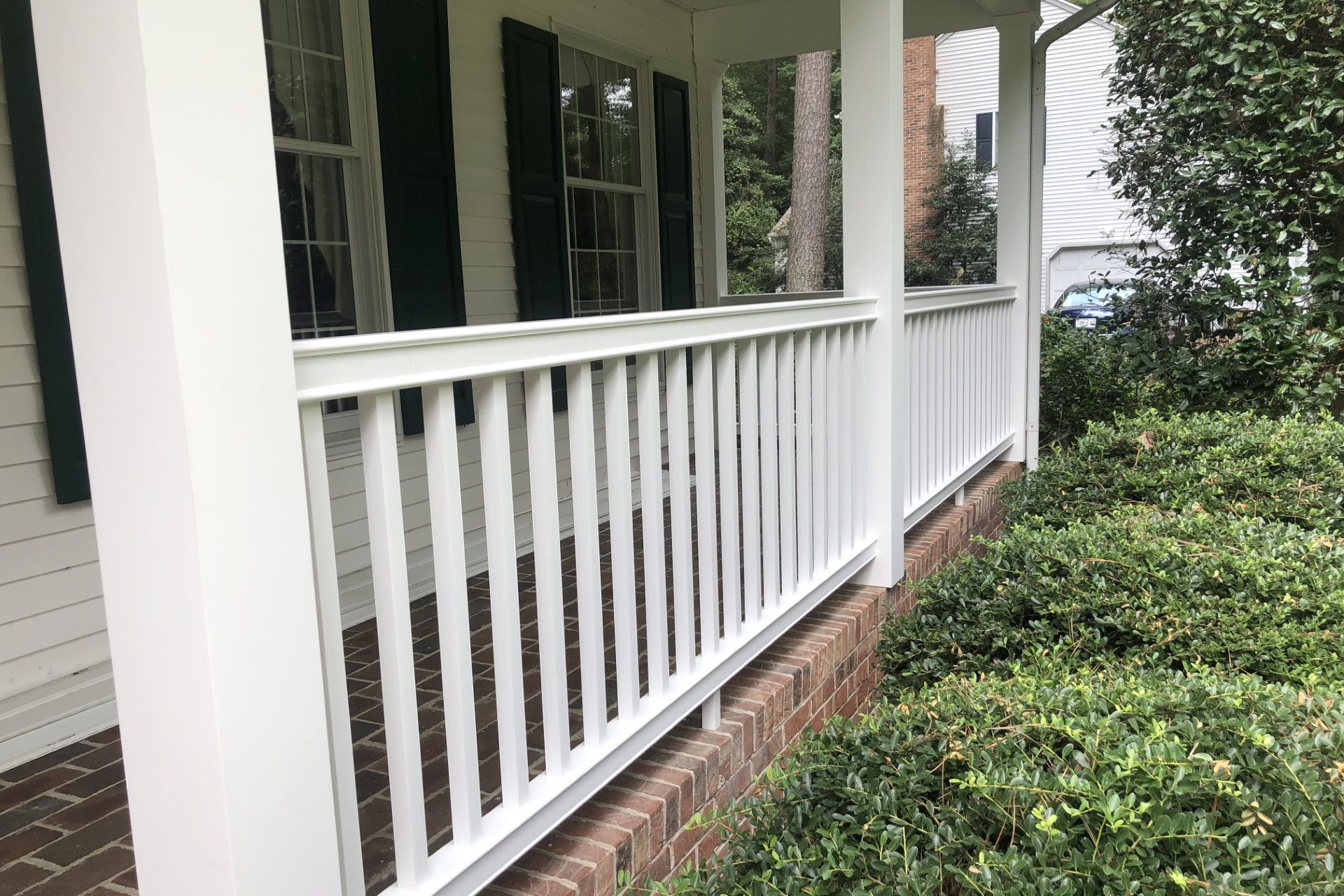 White porch railing with vertical balusters, brick base, and green bushes.