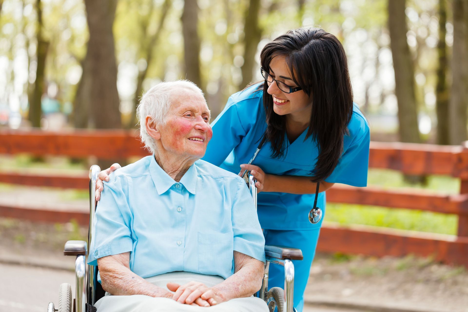 Nurse in blue scrubs smiles at elderly woman in wheelchair outdoors.