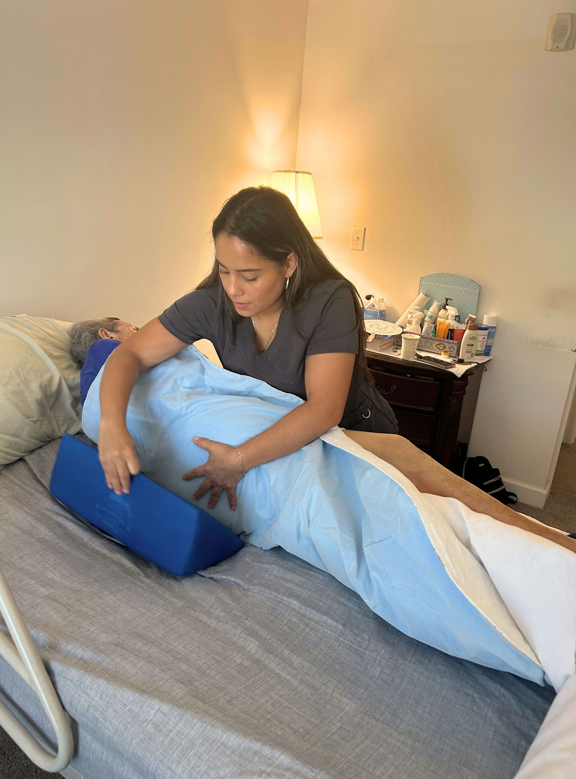 Woman assists an elderly person in bed, using a blue wedge pillow and a blue sheet; bedroom setting.