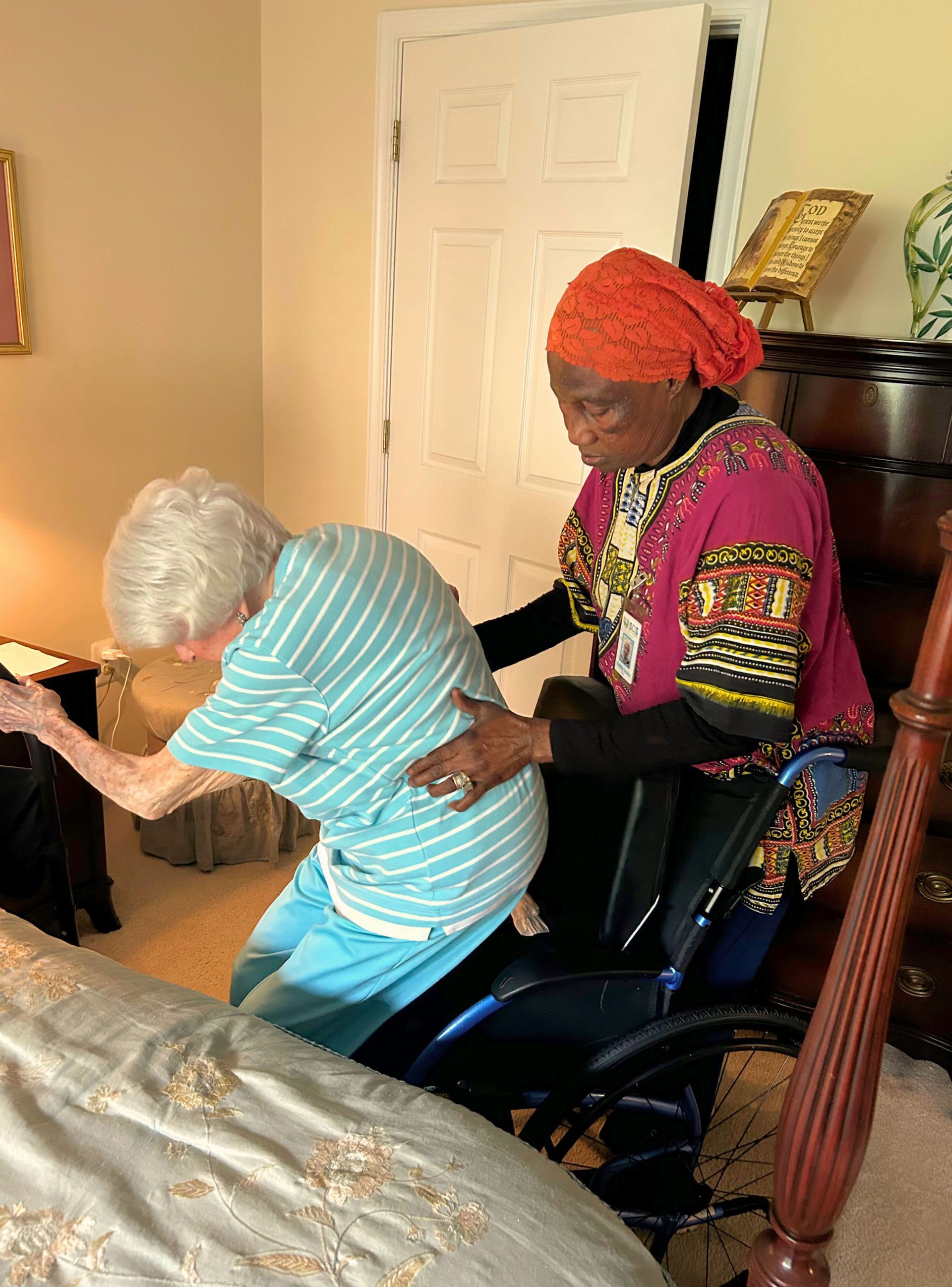 Woman assisting an elderly woman from a wheelchair to a bed, indoors.