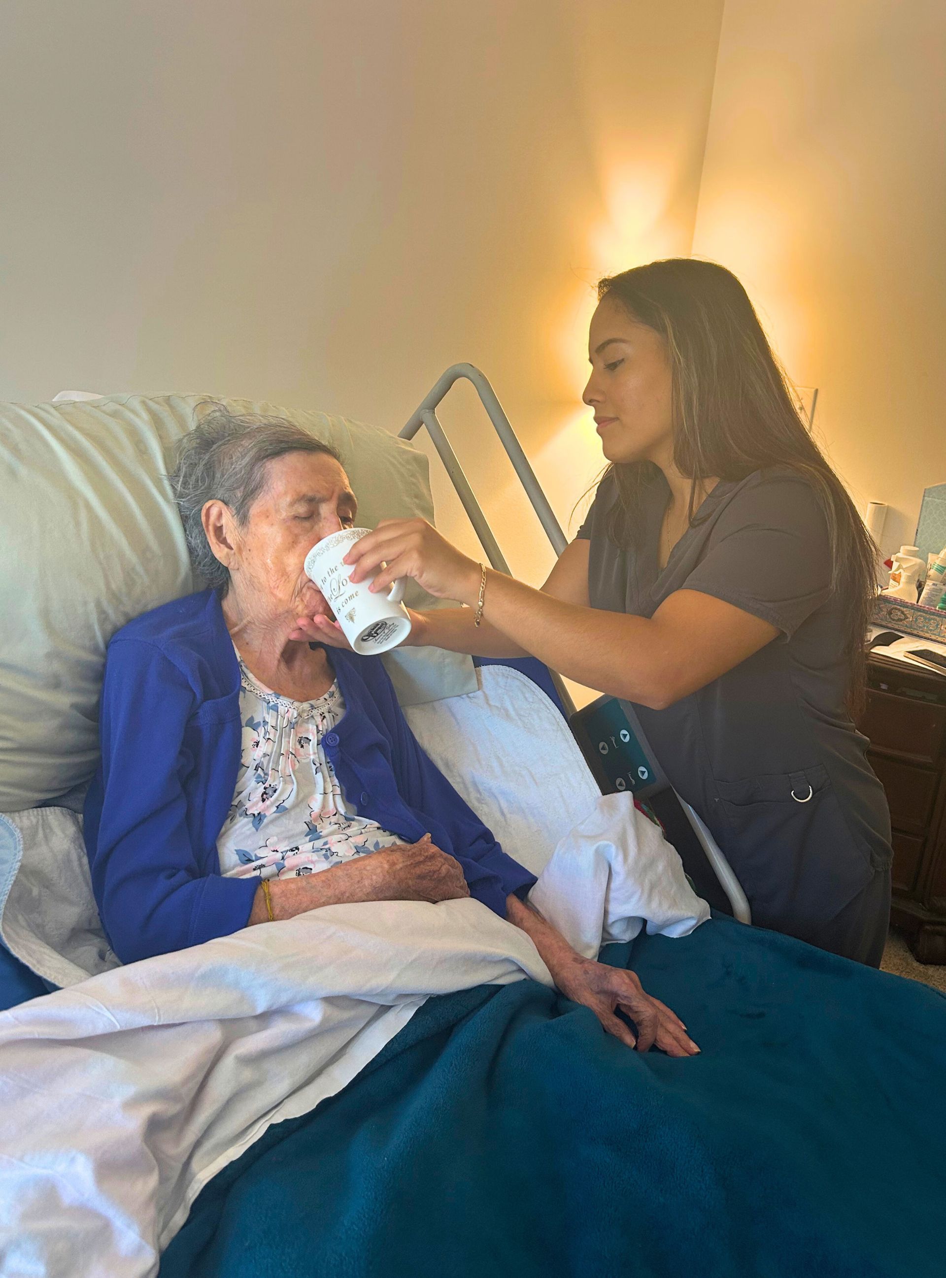 Woman in blue cardigan being helped to drink by a caregiver in a bedroom.