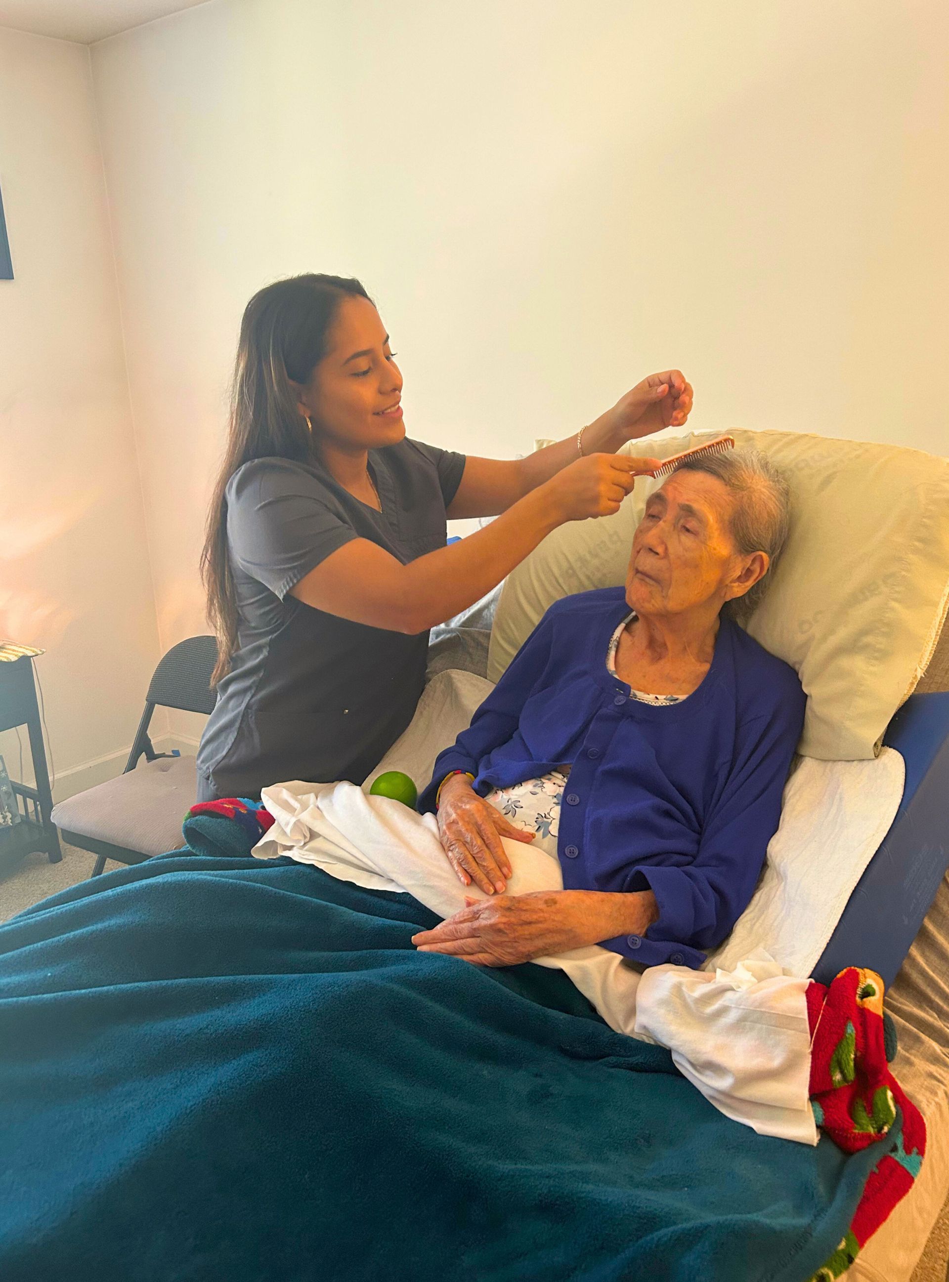 A young woman adjusts an elderly woman's hair while she sits in bed. The woman wears blue.