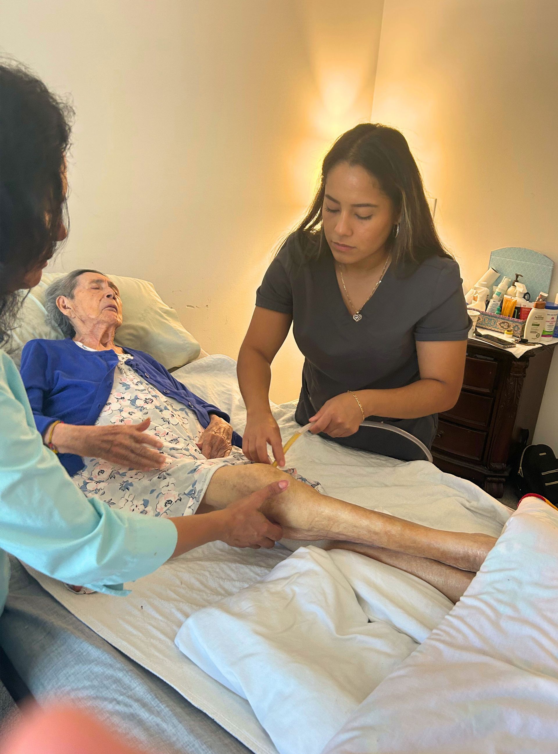 Two caregivers assist an elderly woman on a bed. One massages her leg while the other supports her.