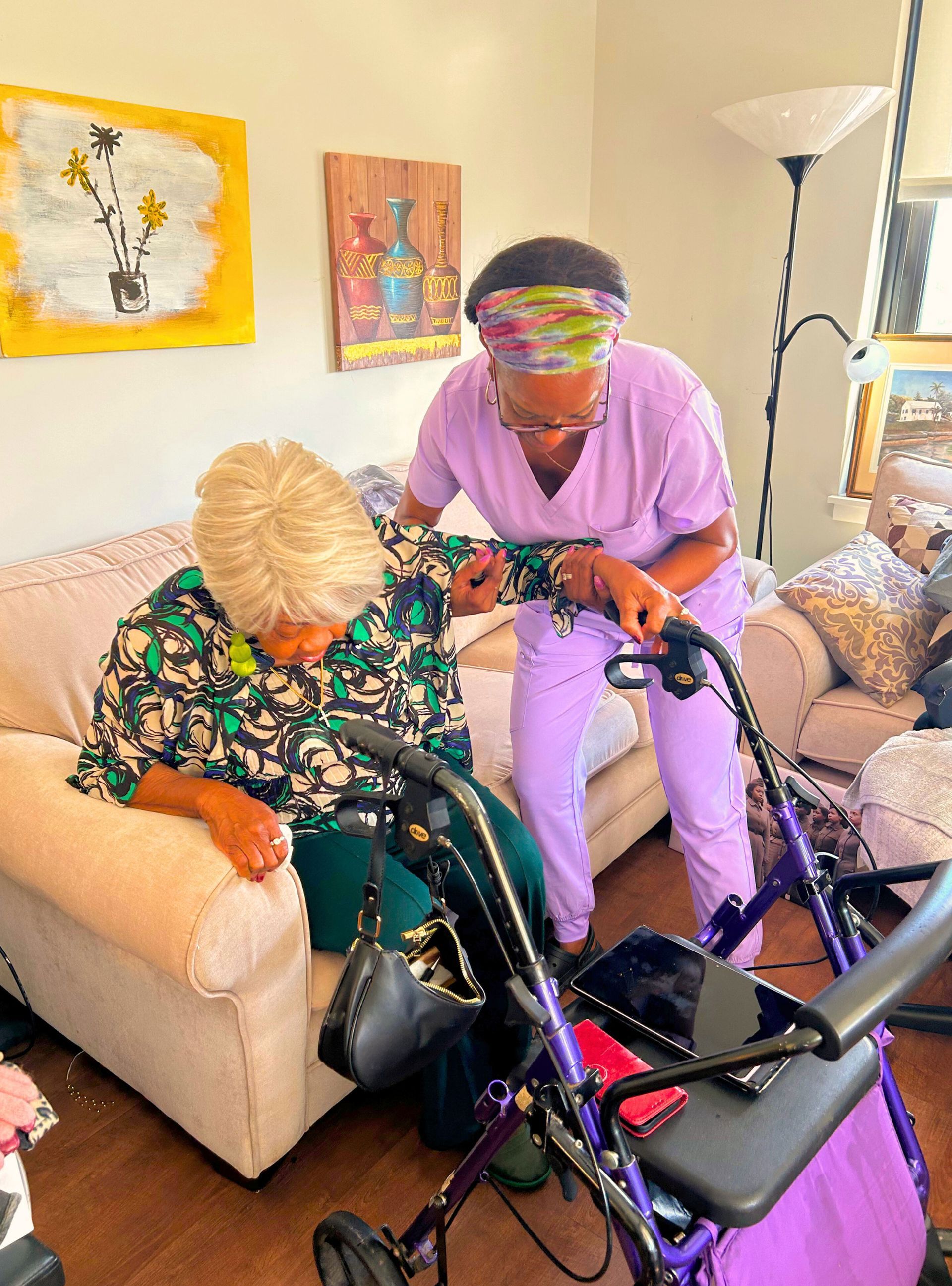 Caregiver assisting elderly woman from a sofa to a walker. Indoor setting with paintings.