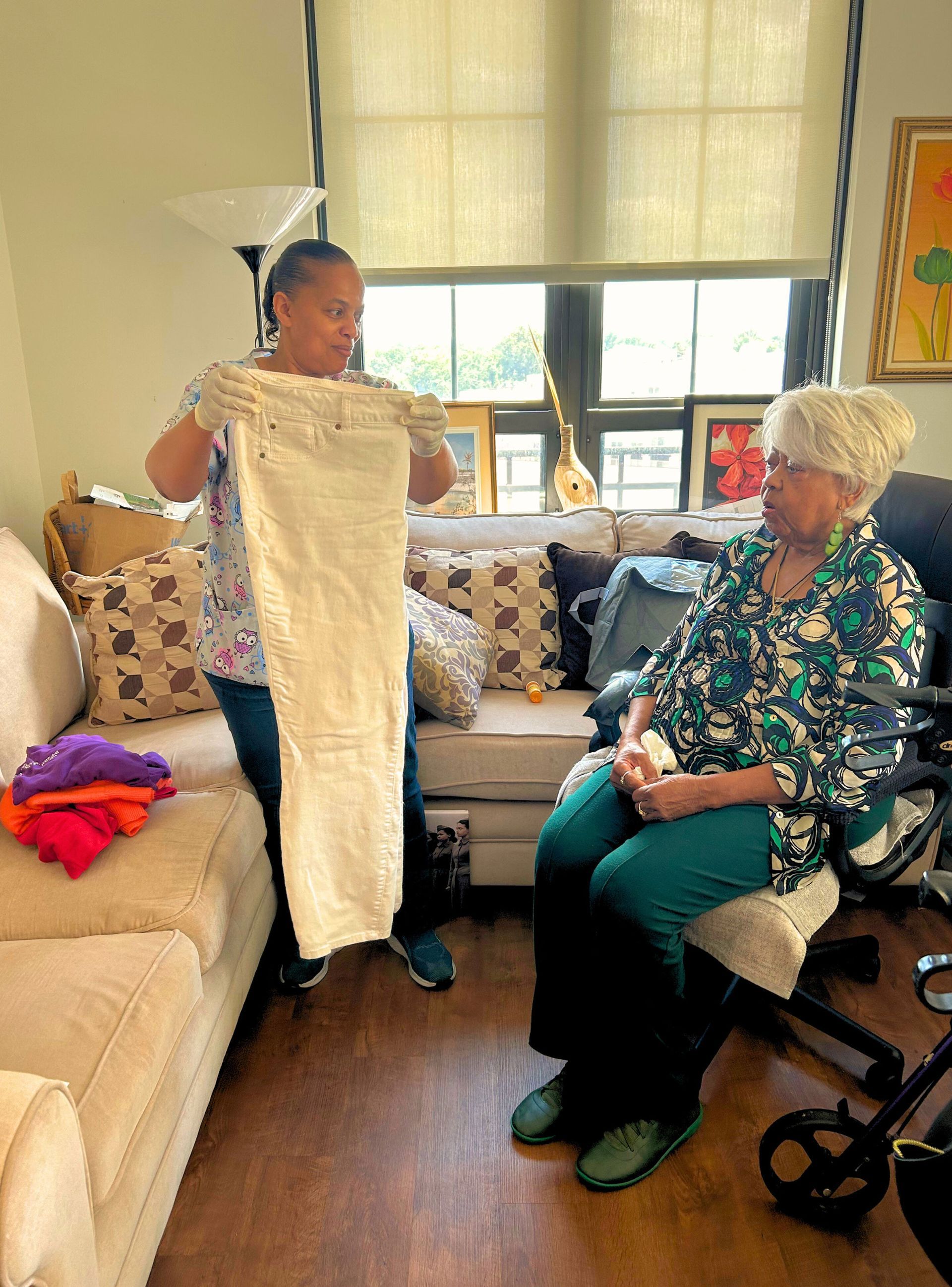 Woman showing off white pants to an older woman seated on a chair, living room setting.