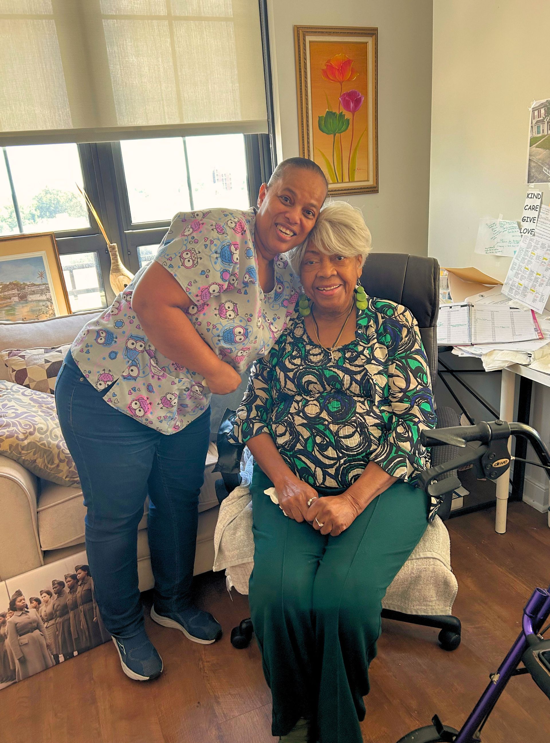 Woman in patterned scrubs hugs an elderly woman in floral top, seated indoors.