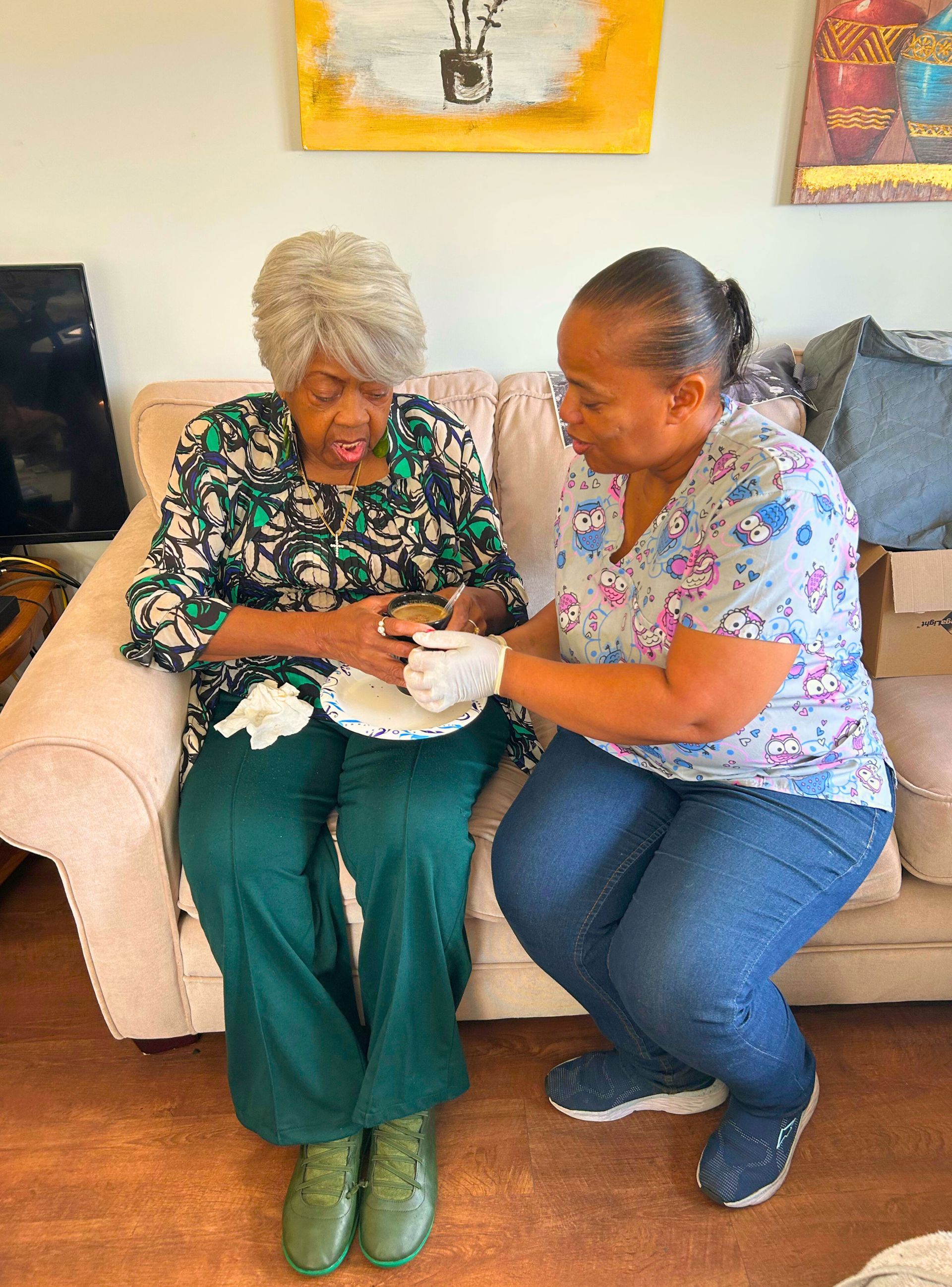 A caregiver assisting an elderly woman seated on a sofa with a snack.