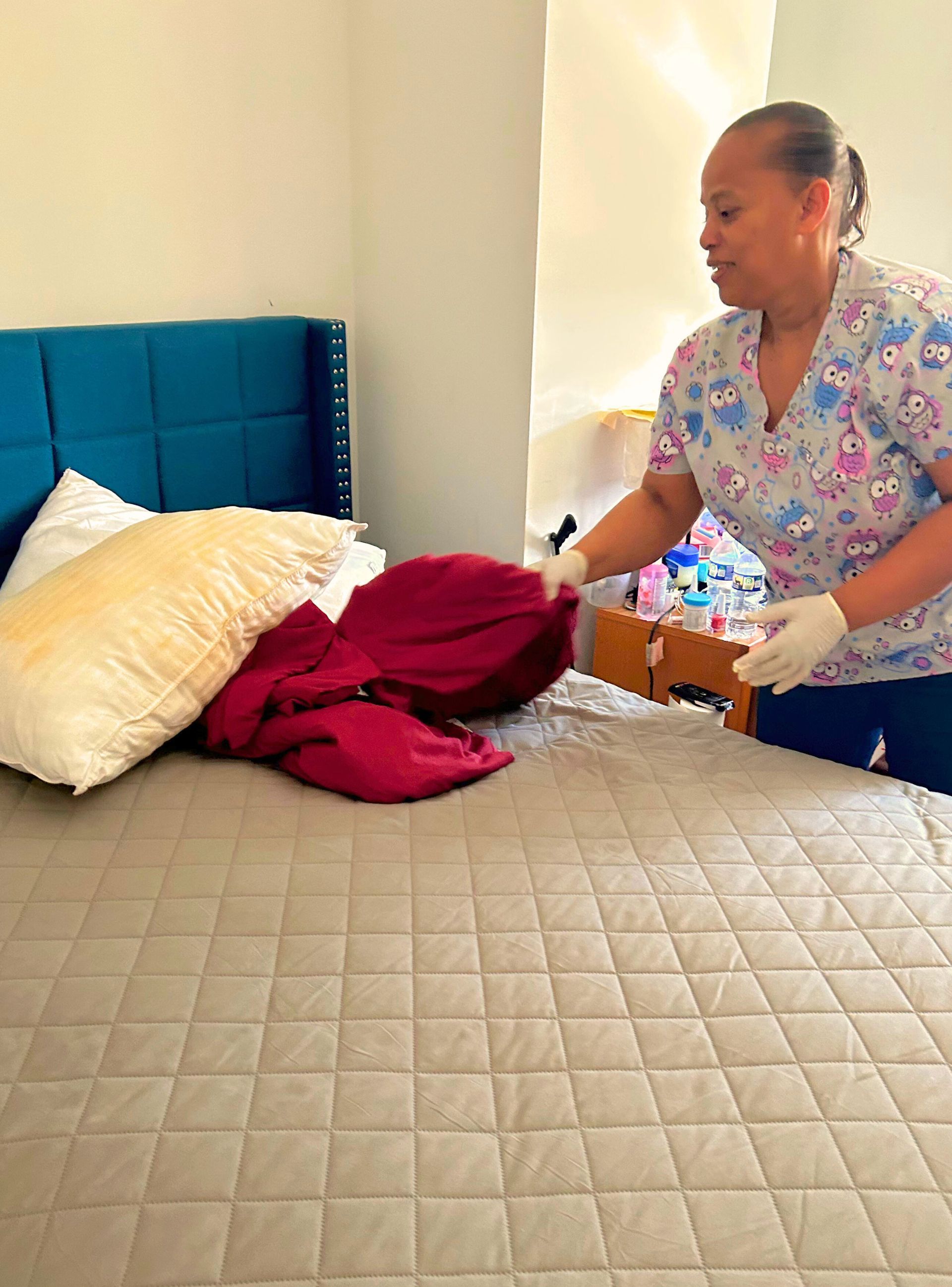 Woman in patterned scrubs makes a bed with a teal headboard, pillows, and red blanket.