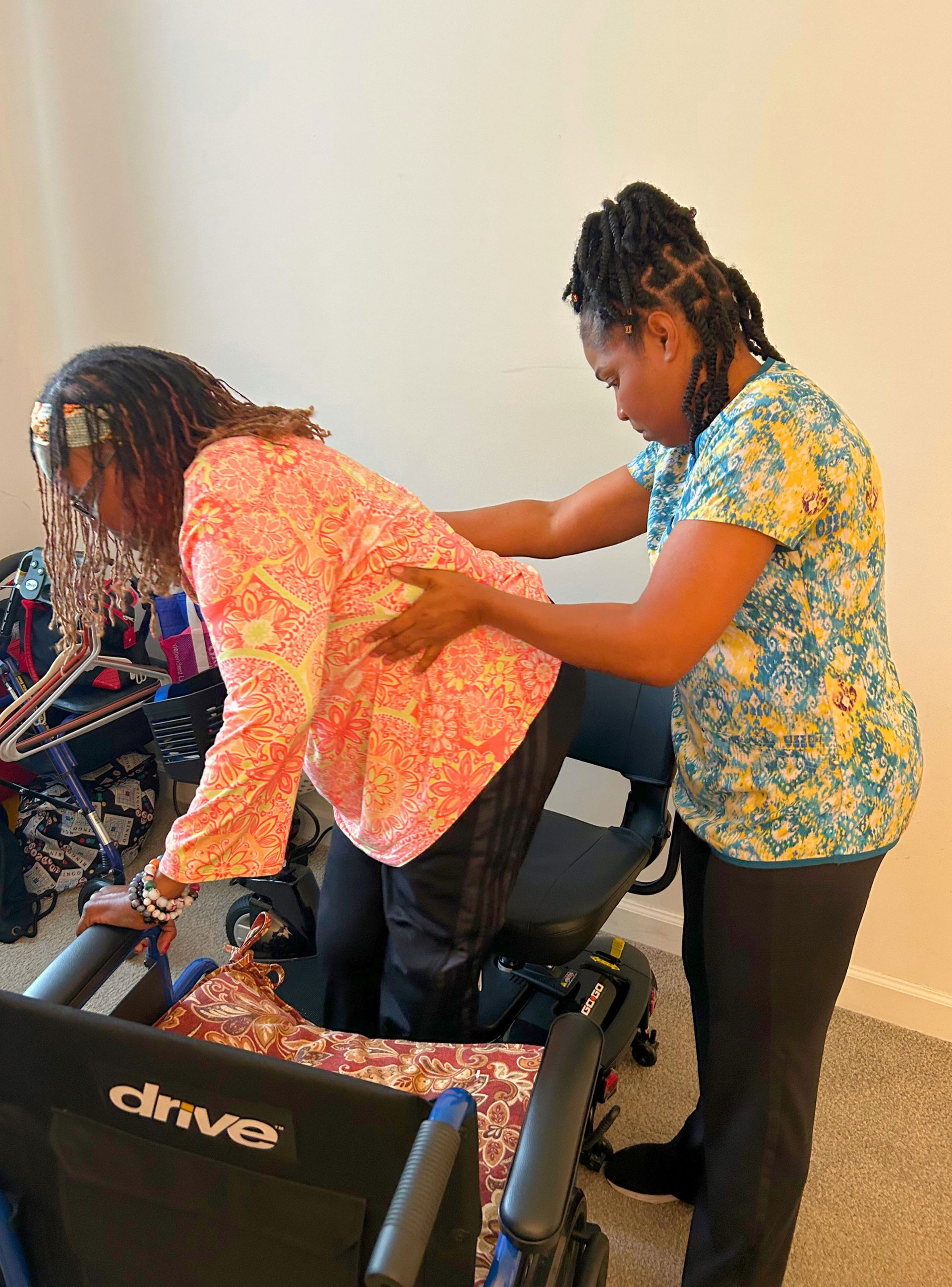 Woman in pink shirt being helped to stand from wheelchair by another woman.