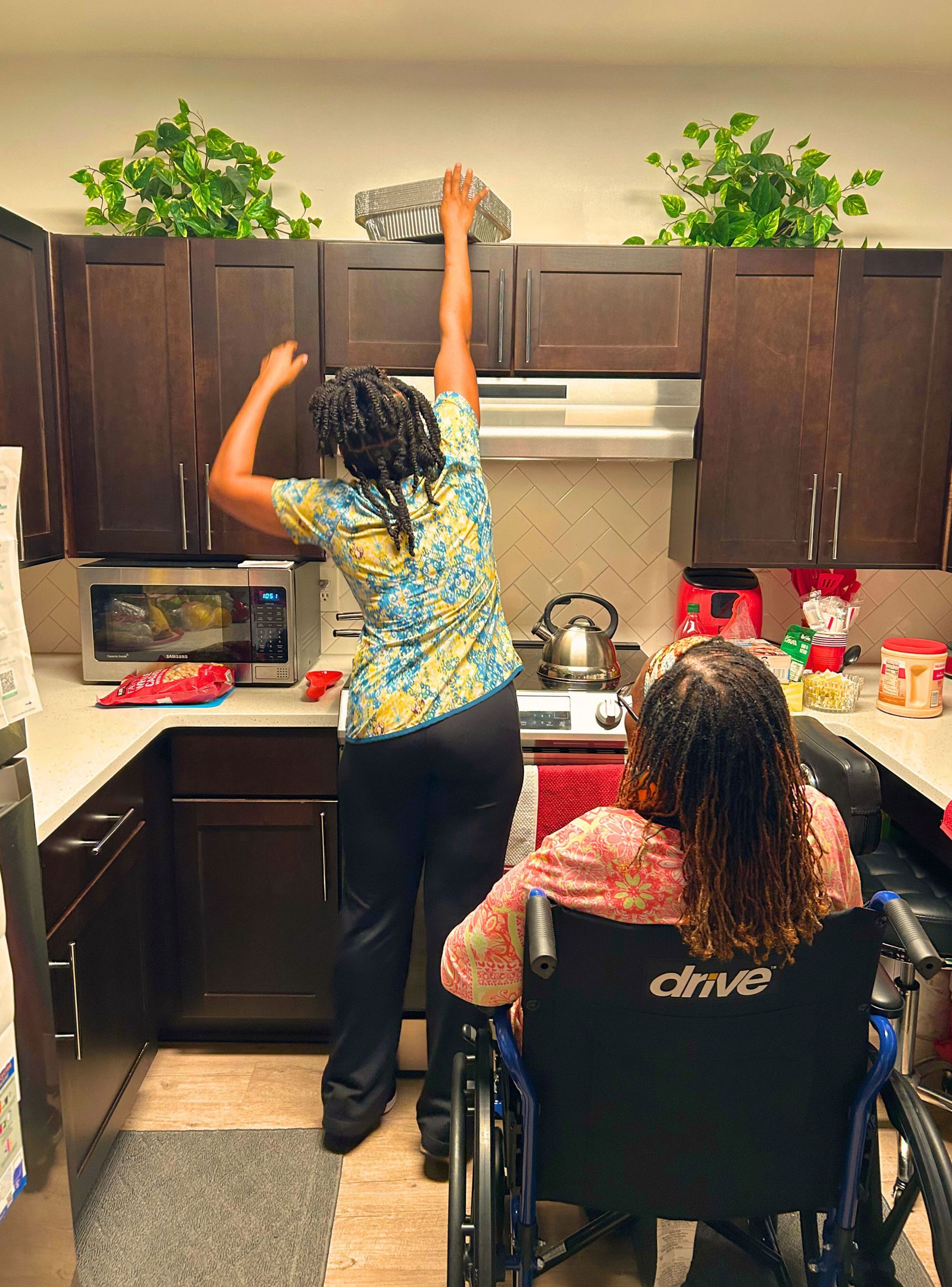 Woman reaching for item on cabinet, another woman in wheelchair watches in kitchen.