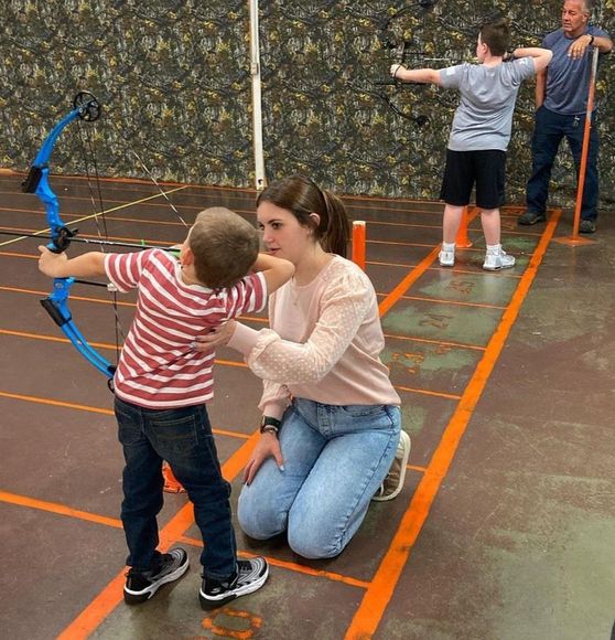 Learn More About Pro Shop Woman assists boy with archery bow in an indoor range. Another person shoots in the background.