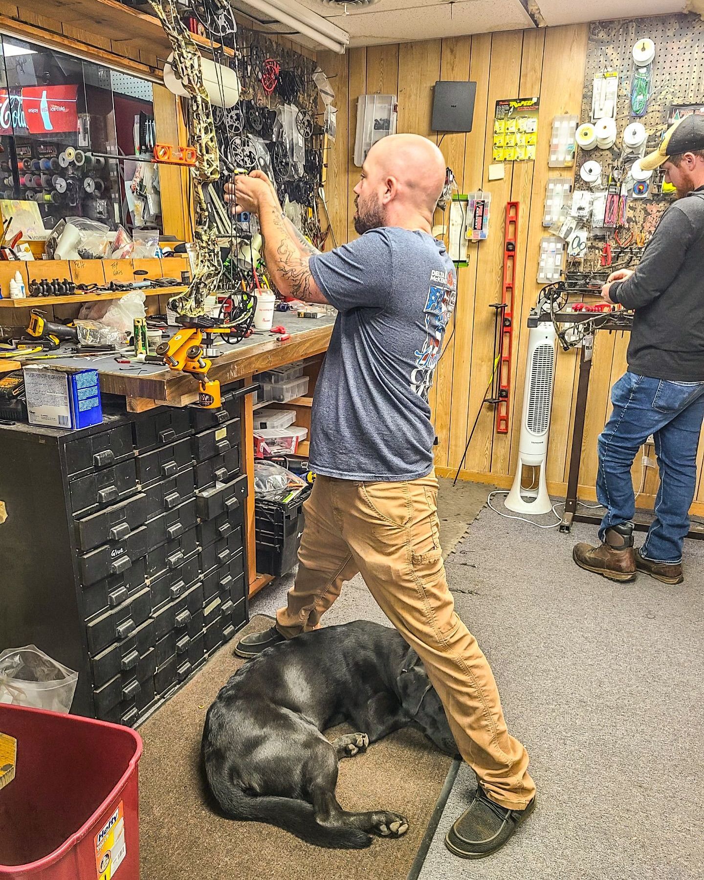 man in shop, standing over dog, working on something. Another man standing nearby.