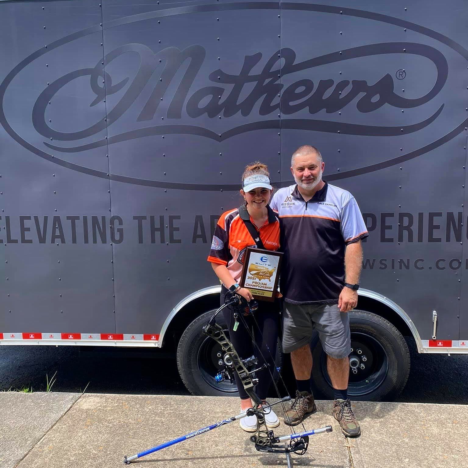 Woman holding trophy with man in front of a Matthews trailer, smiling, bow on ground.