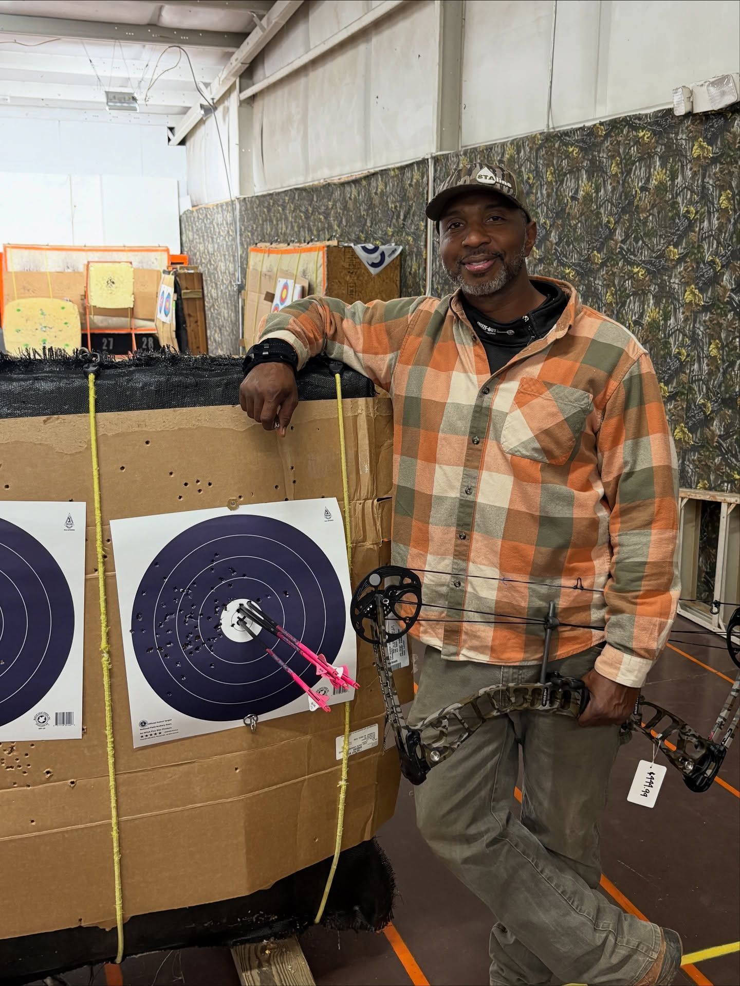 Man leans on archery target, holding bow; indoor range.