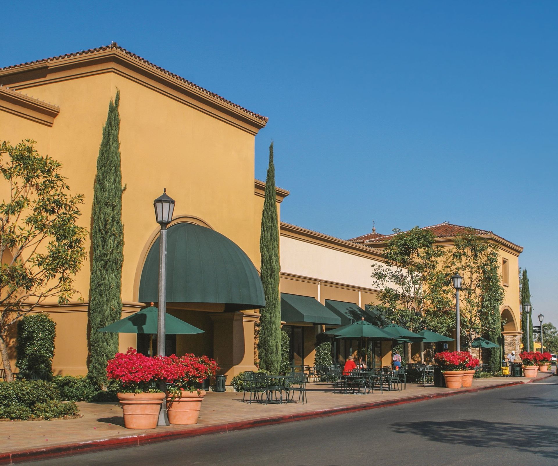 Sunlit shopping center with tan stucco buildings, green awnings, potted flowers, and a clear blue sky