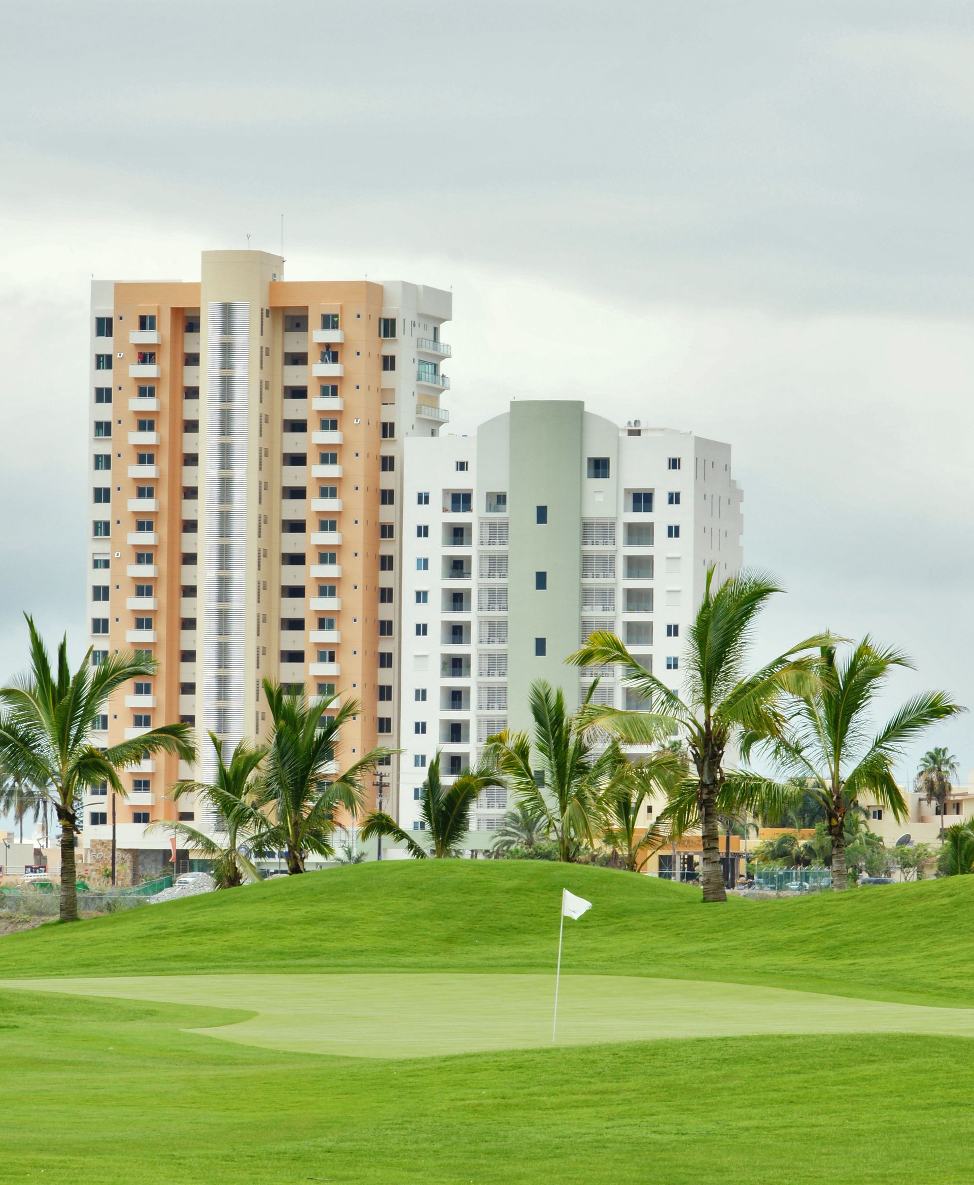 Golf green with a white flag in front of palm trees and tall apartment buildings
