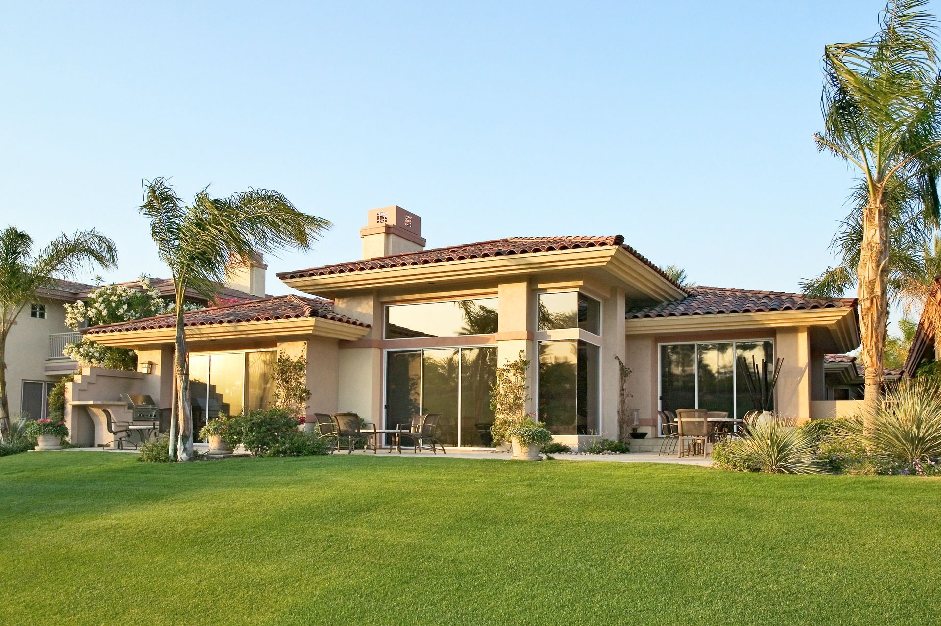 Large beige stucco house with a red-tile roof and green lawn under a clear blue sky