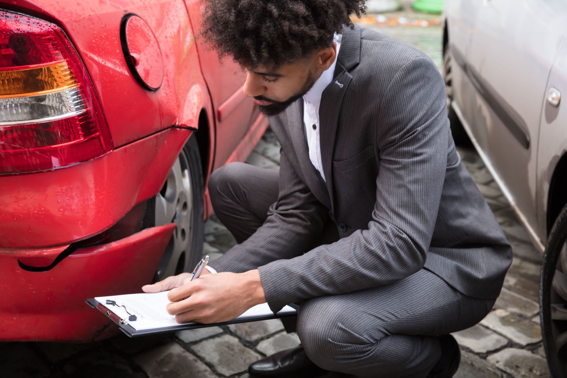 Man crouching beside a red car, writing on a clipboard in a narrow parking area.