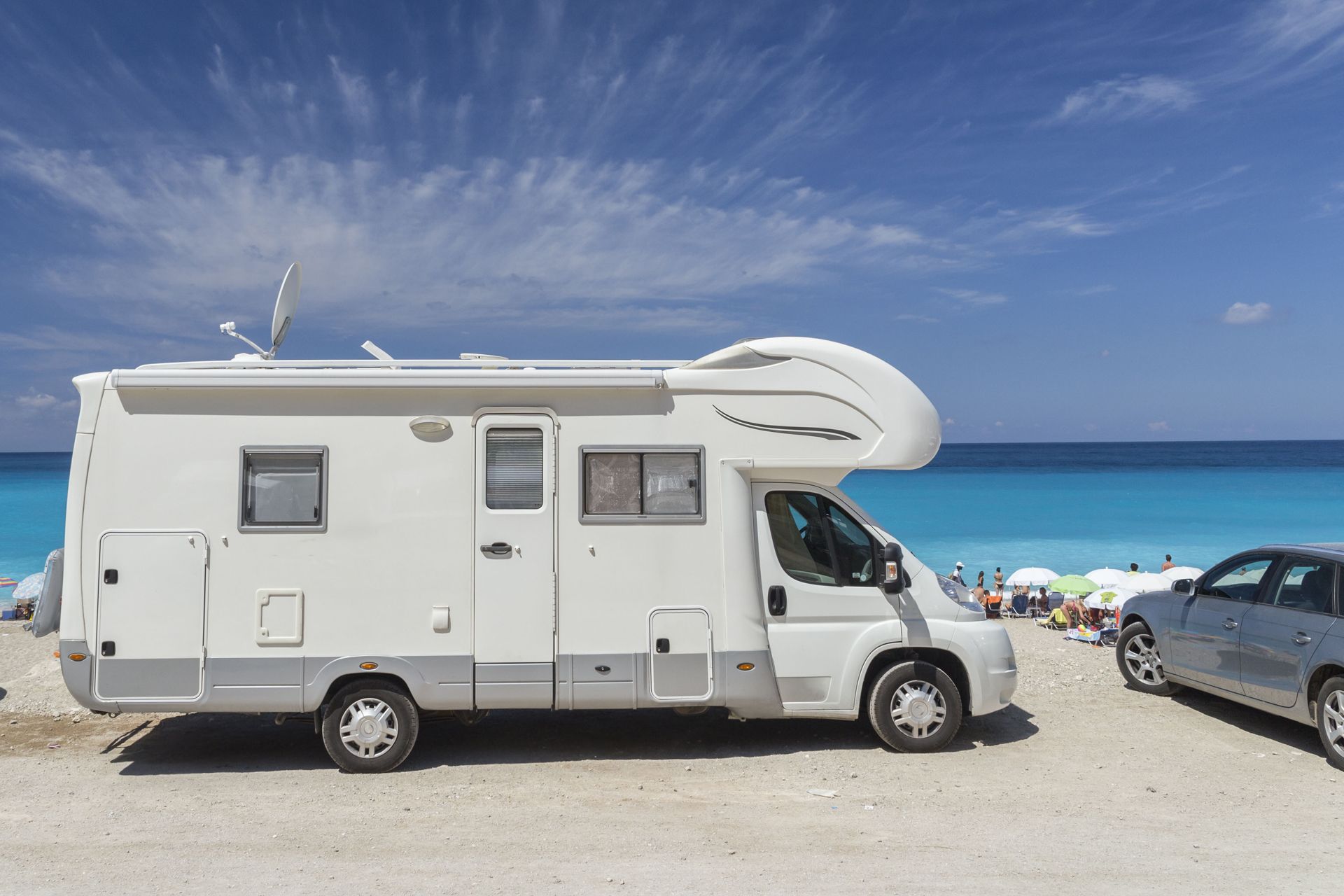 White camper van parked on a sunny beach beside a turquoise sea and another car