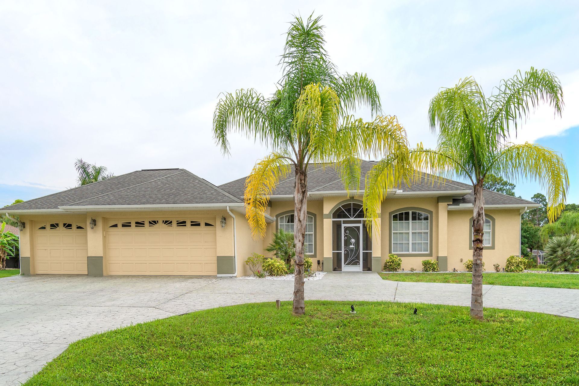 Single-story beige house with palm trees, a two-car garage, and a green lawn under a cloudy sky