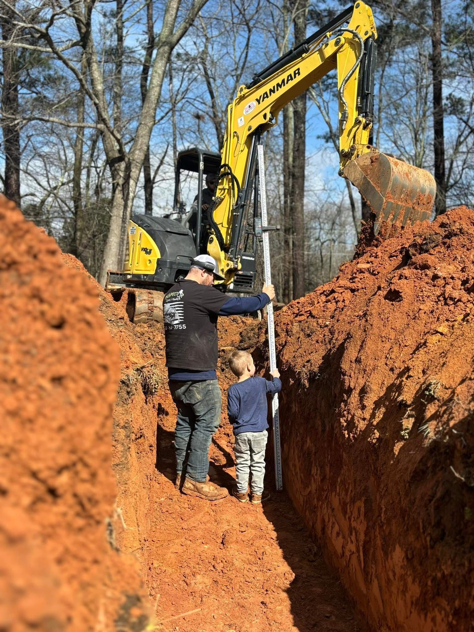 Workers stand in a deep trench as a yellow excavator digs red clay in a wooded area.