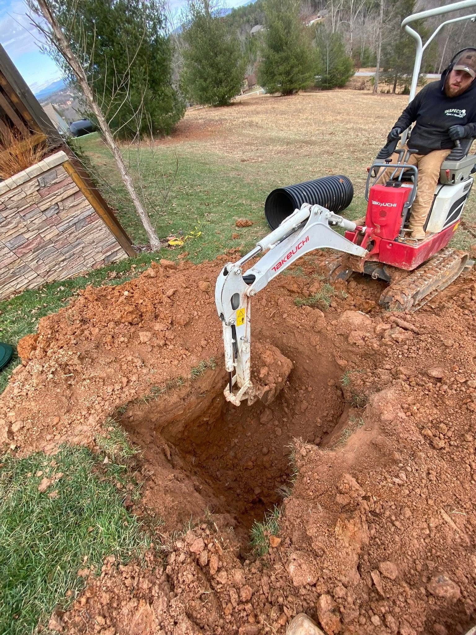 Mini excavator digging a hole in a dirt yard beside a brick wall and trees.