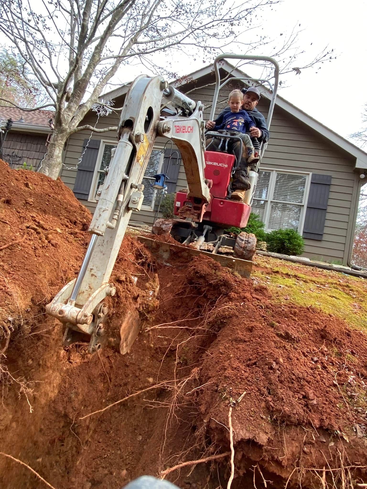 Mini excavator digging a steep dirt trench beside a house, with an operator in the cab.