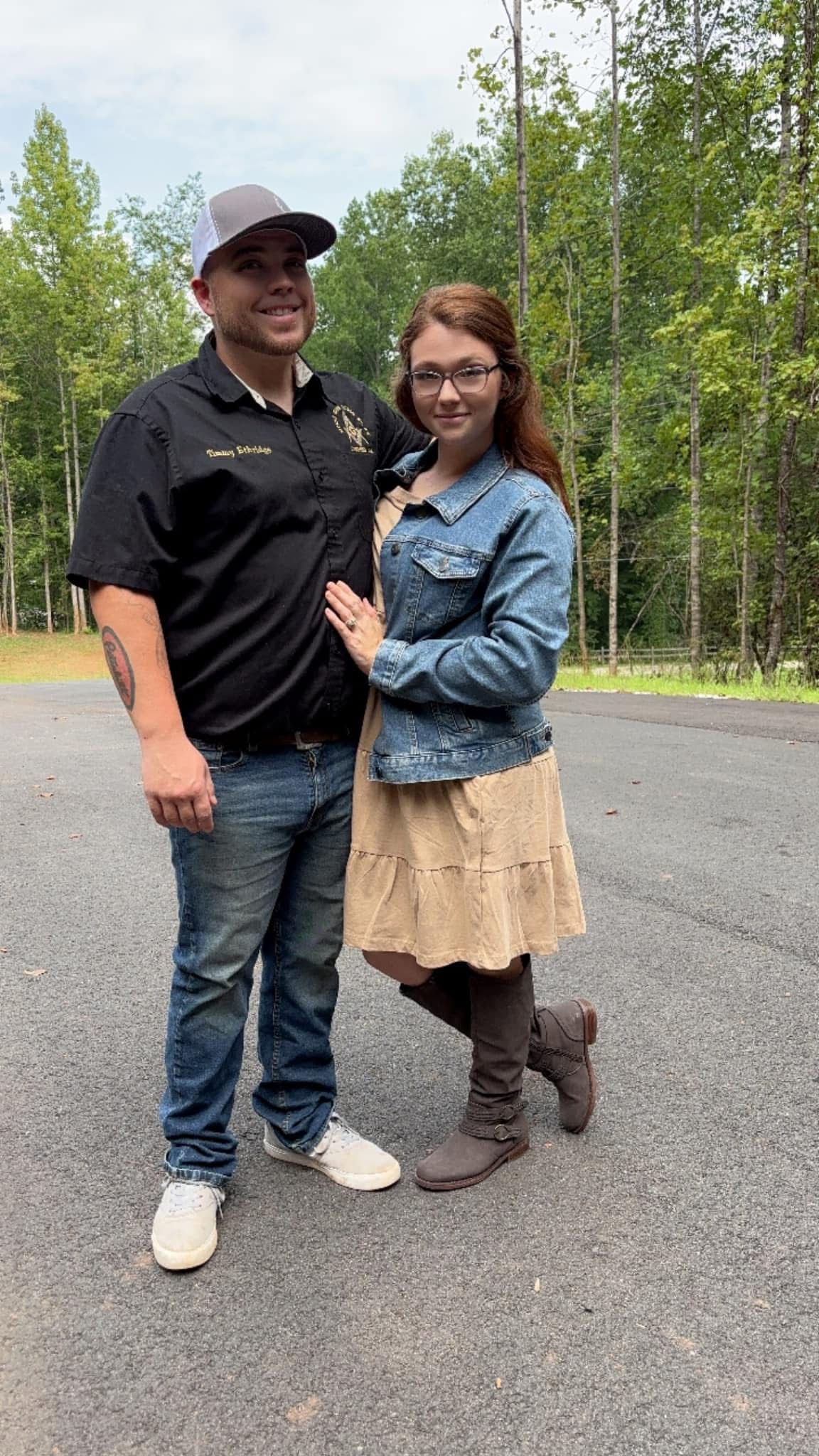 Two people standing together outdoors on a gravel road in a wooded area, smiling at the camera.