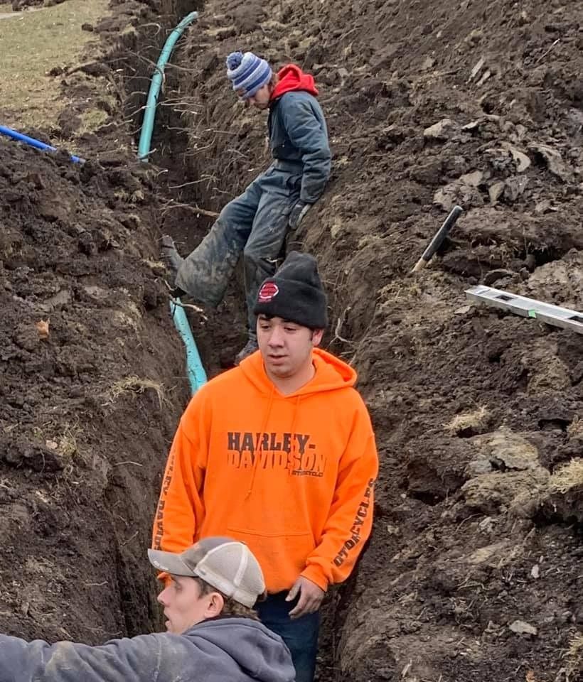 Men working in a trench installing blue pipes; one wears an orange Harley-Davidson hoodie.