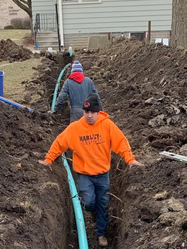 Two workers in a trench installing blue pipe. One in orange hoodie, other in jacket, are surrounded by dirt and an urban setting.