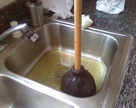 Plunger in a dirty, overflowing kitchen sink filled with murky yellow water.