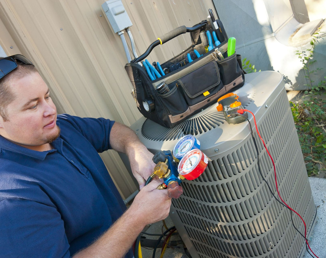 HVAC technician servicing an air conditioning unit outside, using gauges. A toolbox sits nearby.