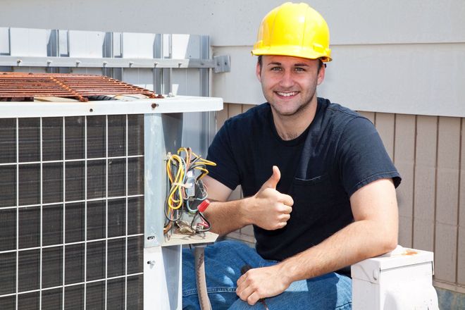 HVAC technician in yellow hard hat giving a thumbs-up next to an air conditioner unit.