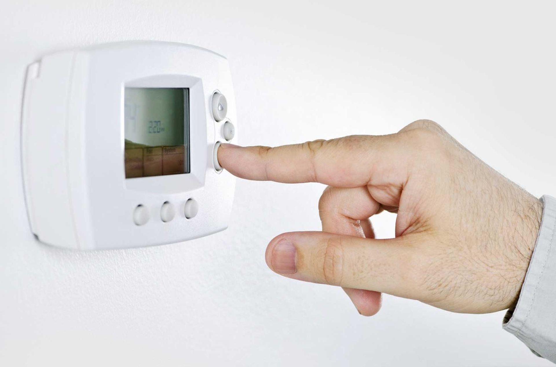 Hand pressing a button on a white thermostat mounted on a white wall.