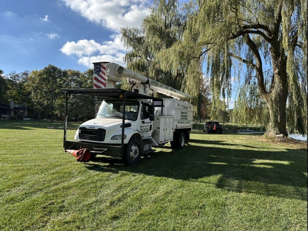 White utility truck with an extended arm, parked on grass near a willow tree, blue sky.