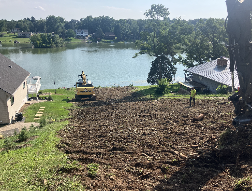Construction site next to a lake; excavator and worker clearing land near buildings and trees.