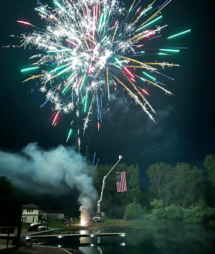 Fireworks exploding over a lake at night, launched from a crane with an American flag.