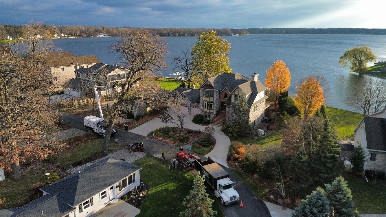 Aerial view of waterfront homes, trees, and lake under cloudy sky. A truck and lift are present for tree work.