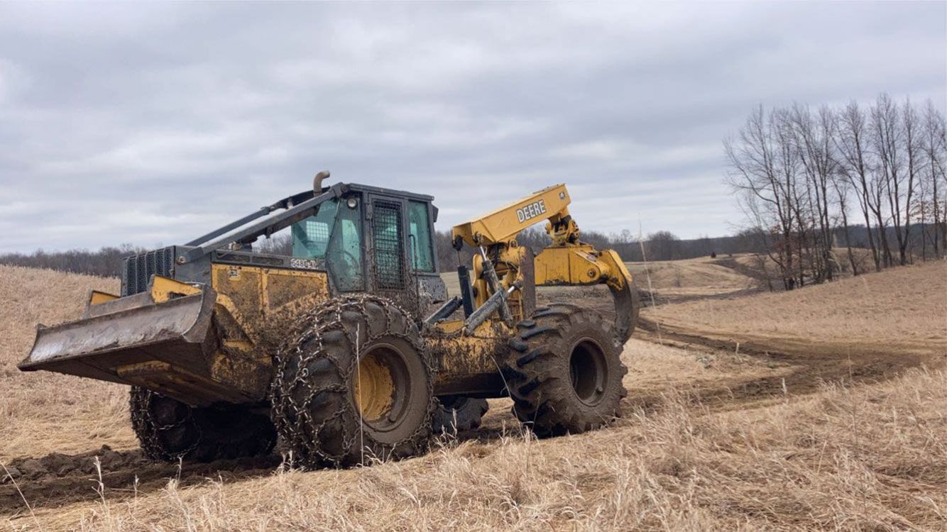 A yellow forestry machine with a blade and log-handling arm on a grassy field; cloudy sky.