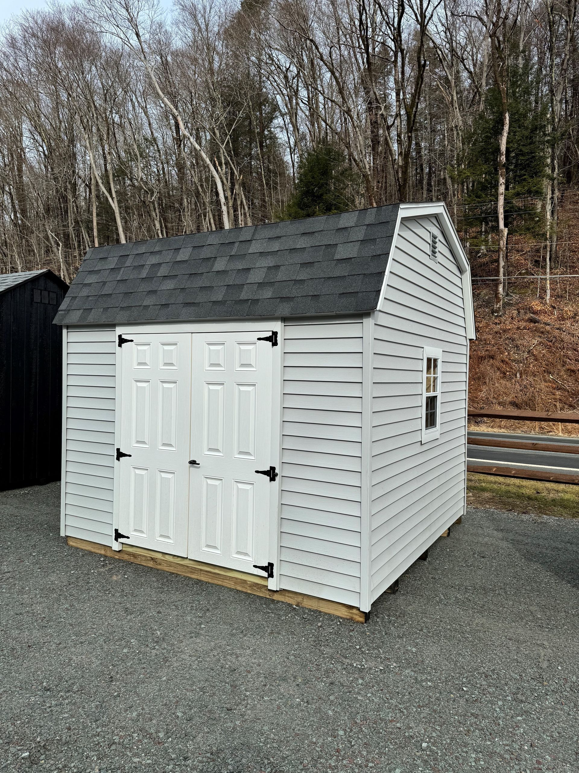 A white shed with a black roof is sitting on top of a gravel lot.