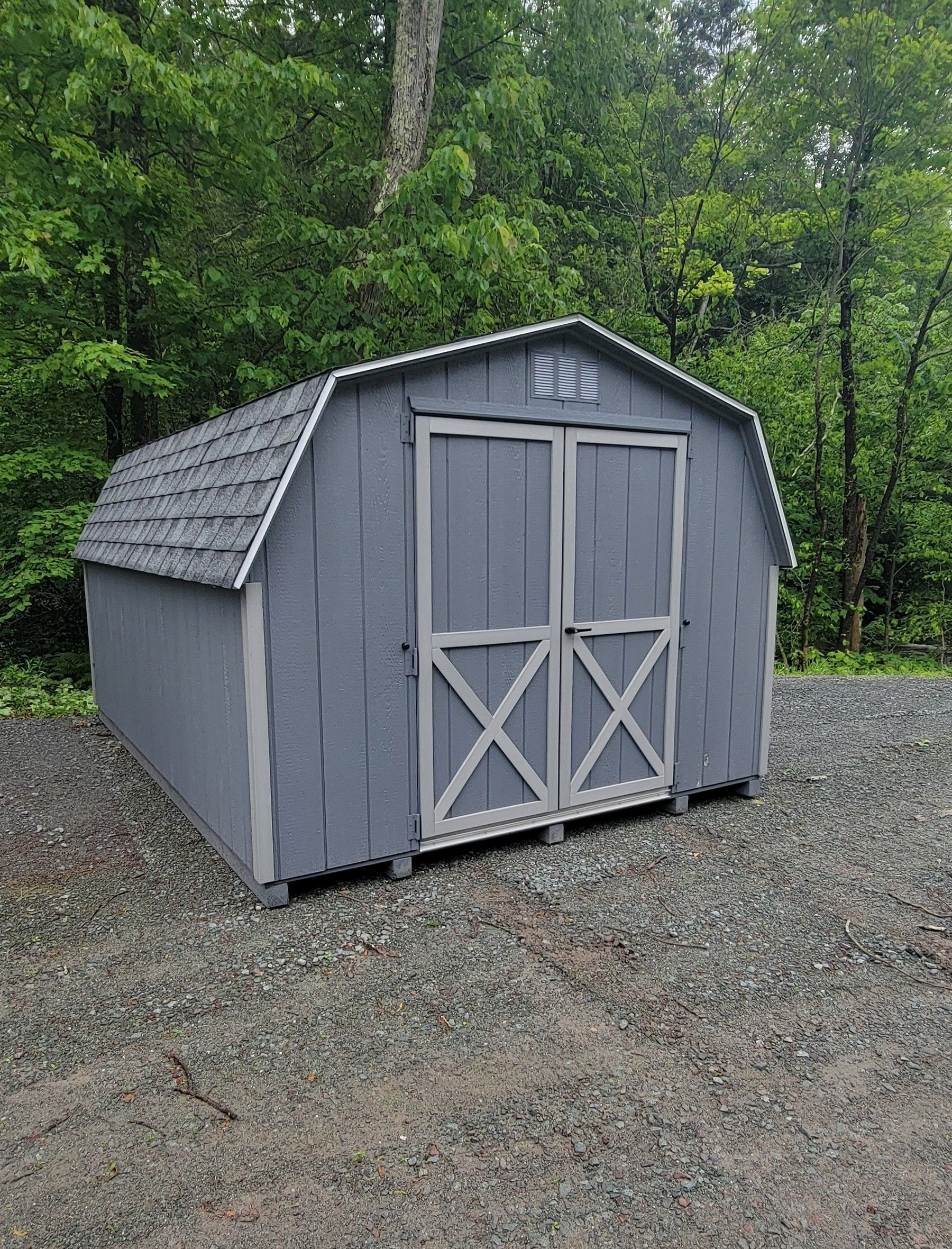 A gray barn shed is sitting on top of a gravel lot in the woods.