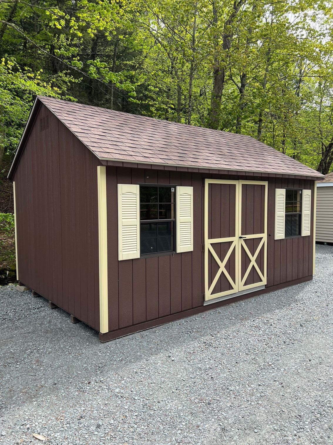 A brown shed with white shutters is sitting on top of a gravel lot.