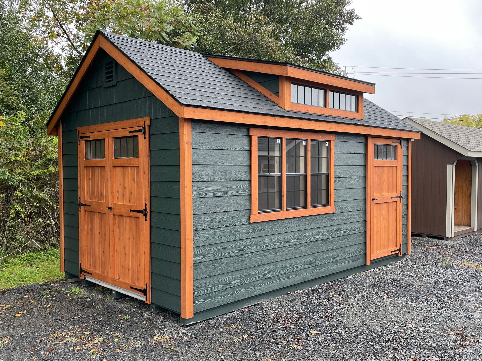 A small green shed with a wooden door and windows is sitting on top of a gravel lot.
