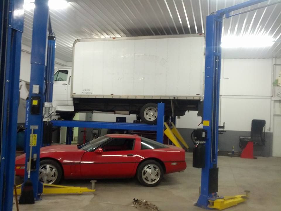 Red car beneath a lifted truck in a garage. Two blue vehicle lifts are visible.