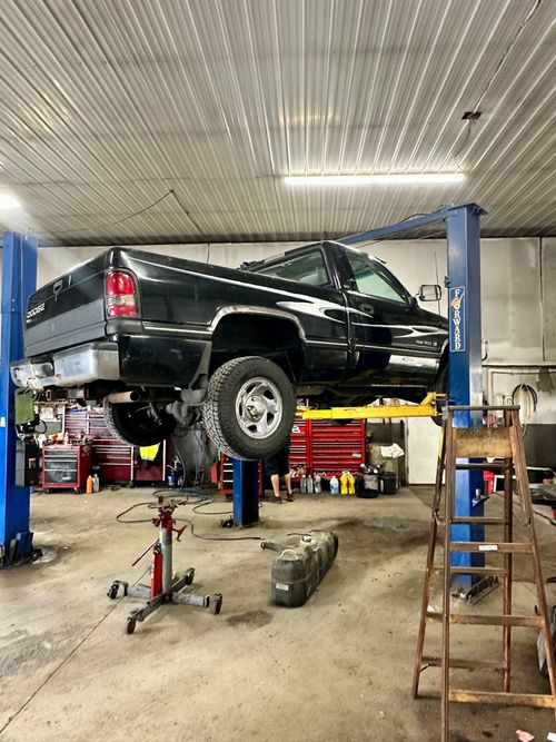 A black pickup truck raised on a two-post lift in an auto repair shop, with a fuel tank resting on the floor below