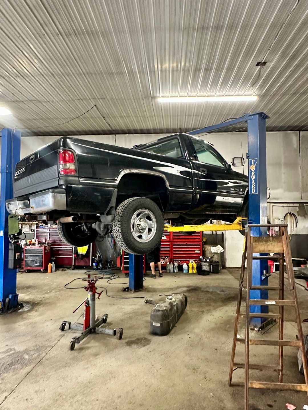 A black pickup truck lifted on a two-post auto lift inside a garage, with a transmission jack and ladder nearby