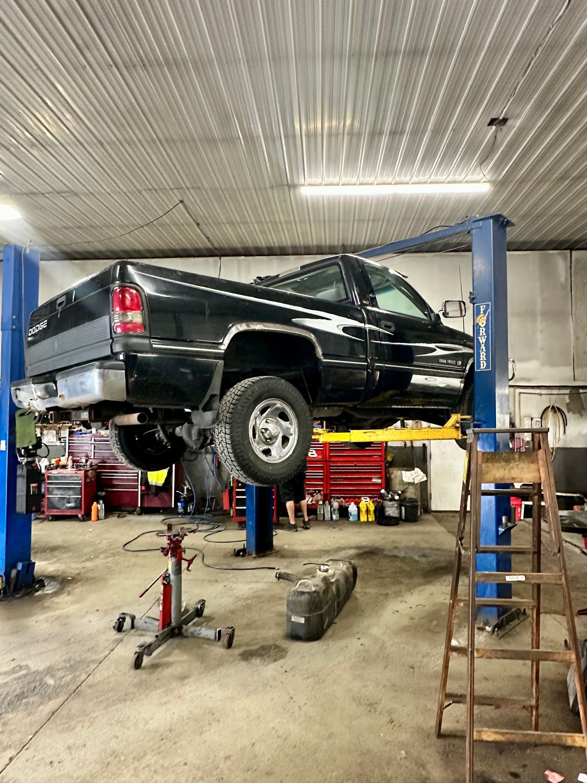 A black pickup truck lifted on a two-post auto lift inside a garage, with a transmission jack and ladder nearby