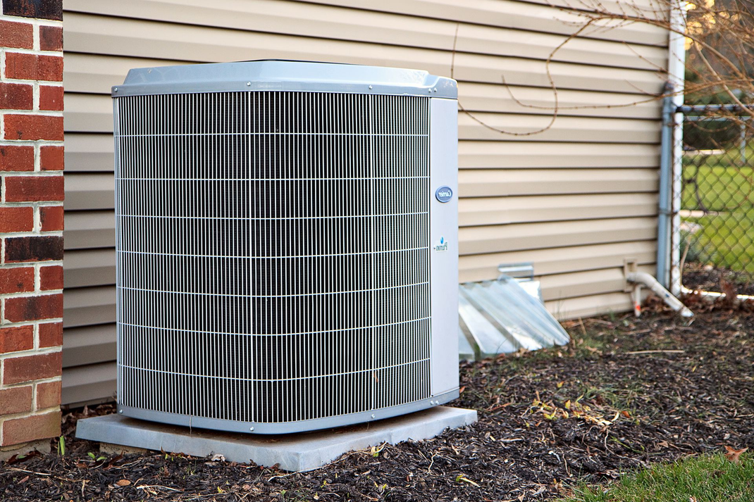 Outdoor air conditioner unit beside a beige house with brick corner and fallen leaves on the ground