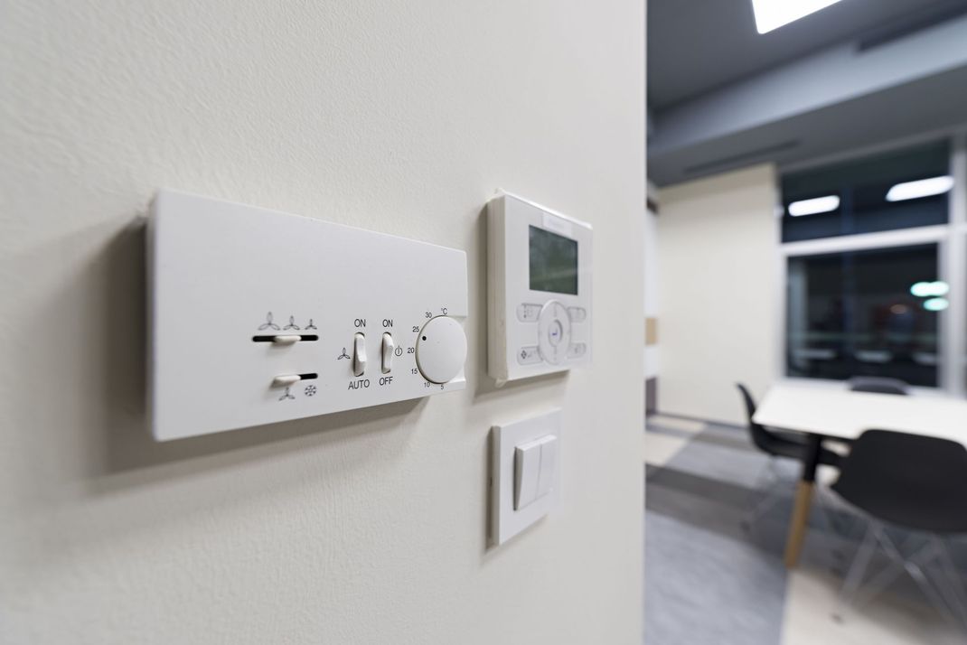 Wall-mounted thermostat controls in a modern office hallway with blurred desks and chairs in the background