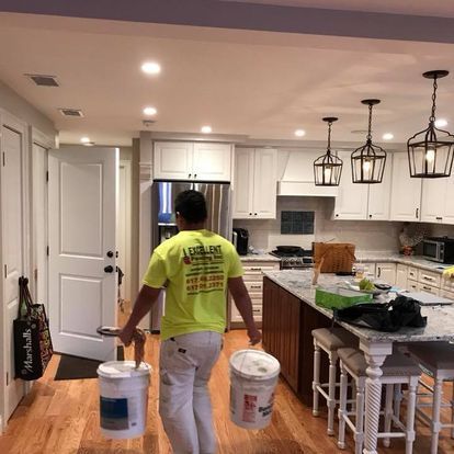 A man in a yellow shirt is carrying two buckets of paint in a kitchen.